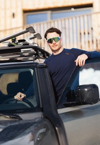 Sort SUV med tagbøjler. Person iført en marineblå langærmet skjorte og reflekterende grønne solbriller, læner sig op ad vinduet, hånden på rattet.