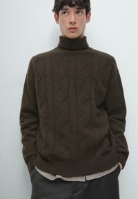 Maglione marrone lavorato a maglia con collo alto, design a trecce, polsini e orlo a coste. Indossato sopra una camicia chiara, abbinato a pantaloni scuri.