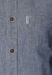 Camisa azul tejida con botones al frente, bolsillo en el pecho, botones marrones y una pequeña etiqueta en el dobladillo. Tela texturizada con un patrón sutil.