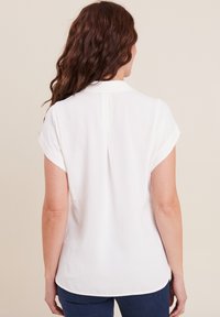 Blouse blanche à manches courtes retroussées, col et pli dans le dos. Le tissu a une texture lisse, avec un ourlet arrondi en bas.