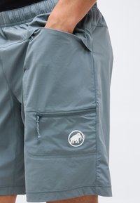 Brazo sin mangas insertado en el bolsillo lateral de unos pantalones cortos grises con cintura elástica, bolsillo con cremallera y logo blanco de mamut en el muslo.