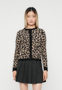 Ung kvinne iført en beige og svart leopardmønstret cardigan med knapper og et mørkegrått plisséskjørt, stående mot en ensfarget bakgrunn.