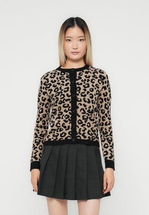 Jonge vrouw in een beige en zwarte panterprint gebuttonde cardigan en een donkergrijze geplisseerde rok, staande tegen een effen achtergrond.