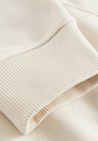 Gros plan d’un poignet en maille côtelée beige sur une manche en tissu doux, montrant la texture et les détails de la couture.