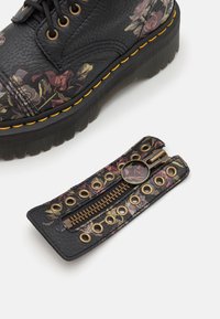 Czarny skórzany but z kwiatowym wzorem, żółtym przeszyciem i czarną podeszwą na platformie. Obejmuje dopasowany kwiatowy dodatek w postaci zamka błyskawicznego.