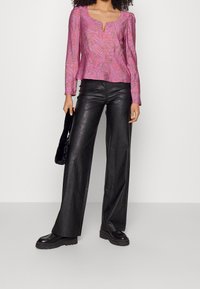 Donna che indossa un top a maniche lunghe rosa con motivo, pantaloni in pelle neri a gamba larga, mocassini neri e tiene una borsa nera in pelliccia.
