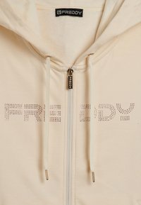 Felpa con zip beige con lettering in strass "FREDDY" sul petto, coulisse abbinate con punte dorate e tirazip con il marchio.