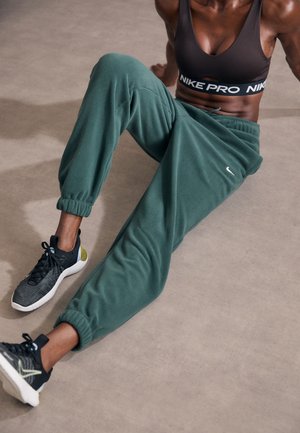 Persona atlética sentada en el suelo, vistiendo un sujetador deportivo oscuro, pantalones deportivos verdes de Nike y zapatillas negras de Nike con suelas blancas y detalles dorados.