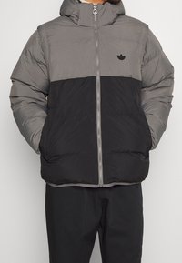 Puffjacka i grått och svart, med dragkedja fram, huva och subtil logotyp på bröstet. Tillverkad av ett texturerat, isolerat material.