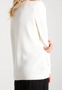 Bílý pletený svetr s texturovaným diamantovým vzorem, žebrovanými manžetami a lemem. Má volný střih a je nošen s černými kalhotami.