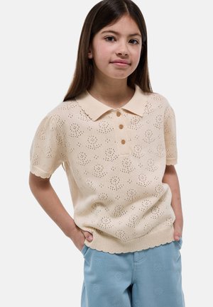 Fille aux longs cheveux bruns portant un haut tricoté beige à manches courtes avec des boutons et un pantalon bleu clair aux motifs floraux subtils, les mains dans les poches.