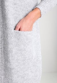 Ljust grå stickad cardigan med en texturerad yta, långa ärmar och en sidoficka med en hand inskjuten i den.