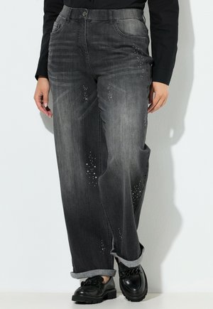 Femme portant un chemisier noir à manches longues, un jean large gris foncé avec de petites taches blanches, et des mocassins noirs ornés d'une chaîne.