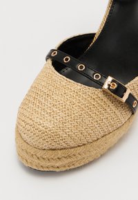 Zapato cuña de yute trenzado con punta redondeada, que cuenta con una correa de cuero negro con detalles en dorado y un acabado natural texturizado.