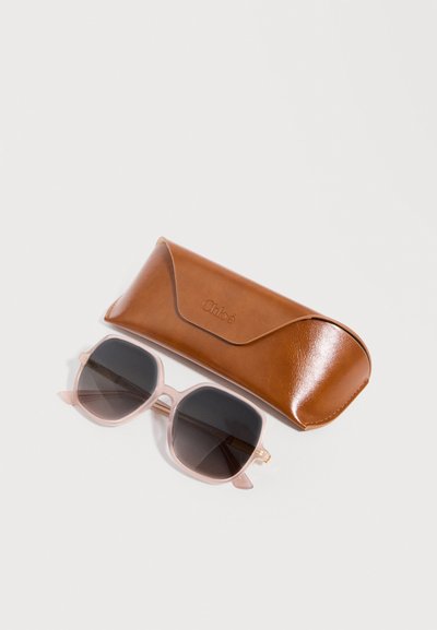Lunettes de soleil à monture carrée avec des cerclages rose clair et des verres sombres, posées à côté d'un étui en cuir marron embossé du logo "Chloé" sur un fond blanc.