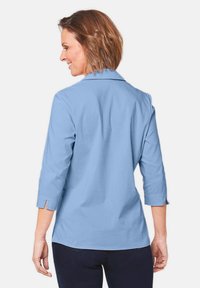 Camicetta azzurro chiaro con un design a collare, maniche a tre quarti e spacchi laterali, realizzata in un tessuto liscio. La vista posteriore mostra una silhouette aderente.