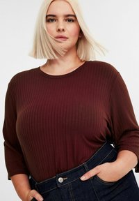 Jonge vrouw met kort blond haar, gekleed in een ribbelige bordeauxrode top en donkerblauwe high-waisted jeans, met haar hand in haar zak.