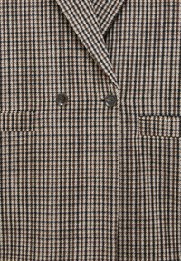 Blazer a motivo houndstooth marrone e crema, doppiopetto, con due bottoni neri e una tasca sul petto sinistro. Tessuto in lana con una texture liscia.