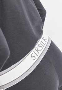 Szare legginsy o gładkiej fakturze, z białym elastycznym pasem w talii, na którym szarym kolorem widnieje nazwa marki "SIKSILK".