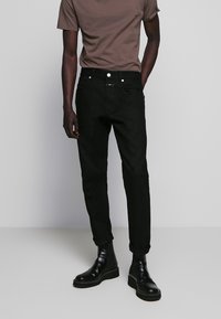Jeans de mezclilla negros con un ajuste slim, diseño de cinco bolsillos y puños enrollados, combinados con una camiseta de cuello redondo marrón y botas negras hasta el tobillo.