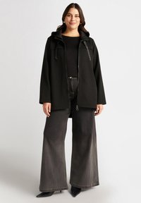 Svart zip-up jacka med huva och dragsko, med en mjuk textur. Kombinerad med vida mörk denimjeans och svarta klackar.