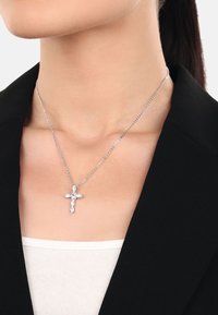 Silverkedja med ett korspendant dekorerat med klara ädelstenar, placerad mot en svart blazer och en vit topp.