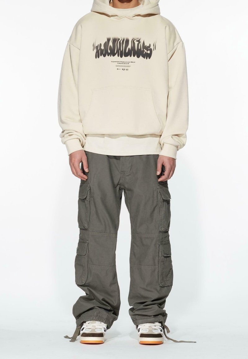 Oversized beige hoodie with graphic print, paired with loose olive green cargo pants featuring multiple pockets and adjustable cuffs.
