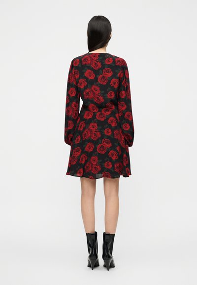 Μαύρο φόρεμα με κόκκινο μοτίβο τριαντάφυλλου, μακριά μανίκια και φαρδιά φούστα. Φορεμένο με μαύρα μποτάκια με ψηλό τακούνι.