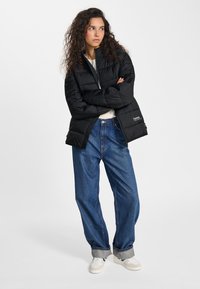 Jonge vrouw met krullend haar, gekleed in een zwarte pufferjas, wit shirt, losse blauwe jeans en witte sneakers, staand met gekruiste armen.