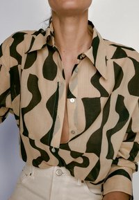 Chemise beige avec des formes abstraites ondulées noires portée sans être rentrée dans un pantalon beige clair à taille haute, laissant apparaître une partie de la poitrine et du cou.