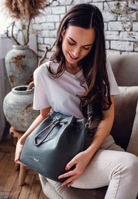 Šedá kožená kabela ve tvaru kyblíku s pleteným popruhem, která má šňůrkové zapínání a kovové akcenty. Hladký povrch a strukturovaný tvar.