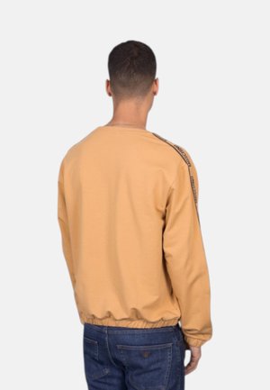 Uomo che indossa una felpa marrone chiaro a maniche lunghe con orlo elastico e jeans blu scuro, di spalle su uno sfondo semplice.