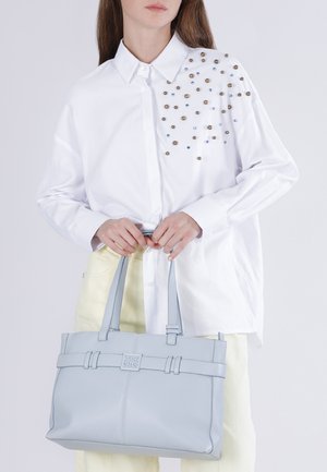 Femme portant une chemise blanche boutonnée avec des clous décoratifs sur l'épaule, tenant un sac fourre-tout en cuir gris clair avec des doubles poignées.