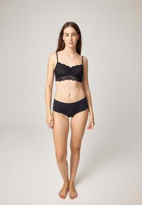 Schwarzes Spitzen-Bralette mit verstellbaren Trägern und einer gezackten Kante. Kombiniert mit einfarbigen schwarzen High-Waist-Slips aus glattem Stoff.