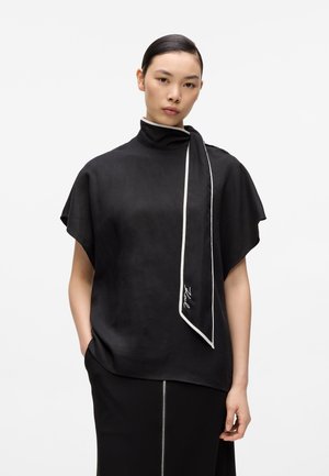 Femeie cu păr neted poartă un top negru cu guler înalt și detaliu de eșarfă cu margine albă și o fustă neagră cu fermoar frontal, stând pe un fundal alb simplu.