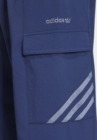 Temno modri Adidas cargo hlače z sprednjim žepom, izvezenim logotipom in tremi odsevnimi črtami ob strani. Gladka tekstura.
