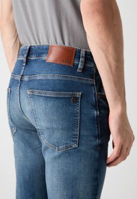 Blugi skinny-fit de denim albastru cu un patch din piele maro la brâu, având buzunare clasice și detalii de cusătură subtile.