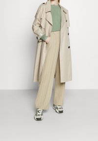 Beige trench coatti mustilla napilla, vihreä neulepaita ja beige raidallinen leveälahkeinen housut, yhdistettynä vihreisiin ja valkoisiin lenkkareihin.