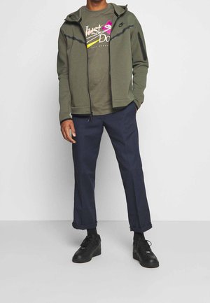 Hombre con chaqueta Nike con cremallera de color verde oliva sobre una camiseta a juego con el lema "Just Do It", pantalones azul marino remangados y zapatillas negras.