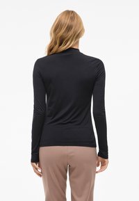 Crni longsleeve top od glatke, rastezljive tkanine. Ima visoki ovratnik i prilagođeni kroj, uparen sa svijetlosmešim hlačama.