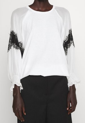 Blouse blanche avec détails en dentelle noire sur le haut des bras, manches longues bouffantes, et poignets à volants, portée avec un pantalon noir.