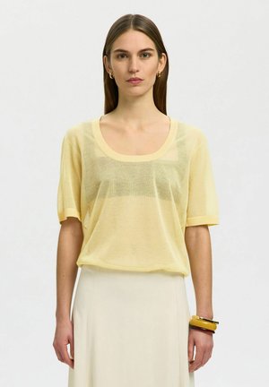 Kvinde med glat brunt hår iført en gennemsigtig gul kortærmet top, cremefarvet nederdel, guld øreringe i ringform og stablede armbånd på højre håndled.