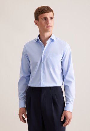 Junger Mann trägt ein hellblaues Hemd mit Brusttasche und dunkle, plissierte Hose, schaut seitlich vor neutralem Hintergrund.