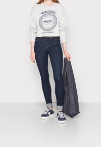 Grijze cropped sweatshirt met marineblauwe grafiek, donkerblauwe skinny jeans en marineblauwe sneakers. Houdt een donkergrijze totebag vast. Soepele texturen overal.