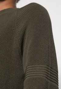 Das olivgrüne Strickpullover zeichnet sich durch ein strukturiertes Rippenmuster, geschichtete Maschendetails an den Schultern und ein lockeres Design aus.