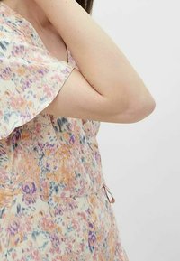 Haut du corps d'une femme en robe à manches courtes florale avec un motif pastel orange, violet et bleu, bras fléchi avec la main près du cou contre un fond uni.