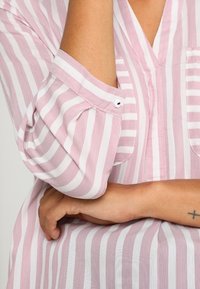 Camicia a righe rosa chiaro e bianca con scollatura a V. Le maniche sono arrotolate e dotate di bottoni. Tessuto morbido e testurizzato.