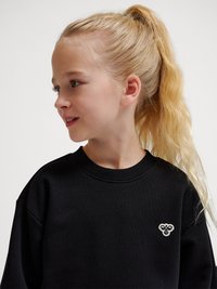 Lyshåret pige med hestehale iført en sort sweatshirt med et lille hvidt emblem på venstre bryst, kigger til siden.