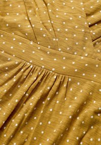 Musztardowa sukienka w białe kropki, z teksturowanej tkaniny, z marszczoną talią i plisowanymi detalami. Gładka i lekka.