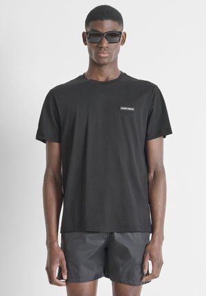 Joven con gafas de sol negras, camiseta negra con pequeño logo y pantalones cortos oscuros, de pie frente a un fondo liso y claro.